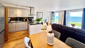 A modern kitchen with a dining area, a sofa, and a view of the sea.