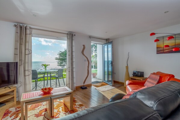 A modern living room with a large window, a red sofa, a TV, and a dining area with a view of the sea.