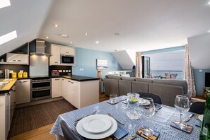Een moderne keuken met inbouwapparatuur, een eethoek met een tafel gedekt voor twee personen, en uitzicht op zee door grote ramen.