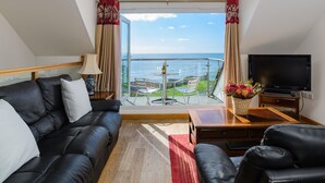 Un soggiorno moderno con un divano in pelle nera, un tavolino in legno e una televisione a schermo piatto. Un balcone con ringhiera in vetro si affaccia sul mare.