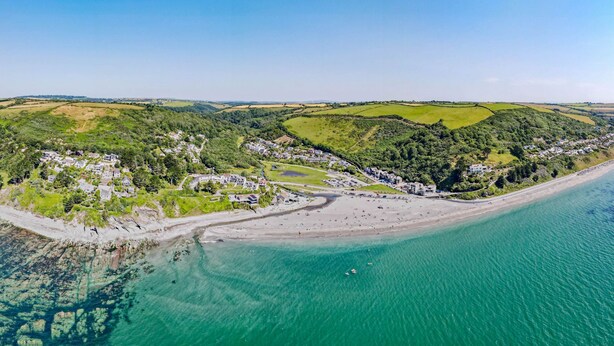 An aerial view of a coastal town with a beach, green hills, and residential areas.