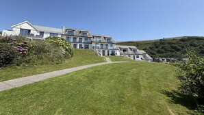 A modern multi-story building with a sloped roof, a paved pathway leading to the entrance, and a lush green landscape in the background.