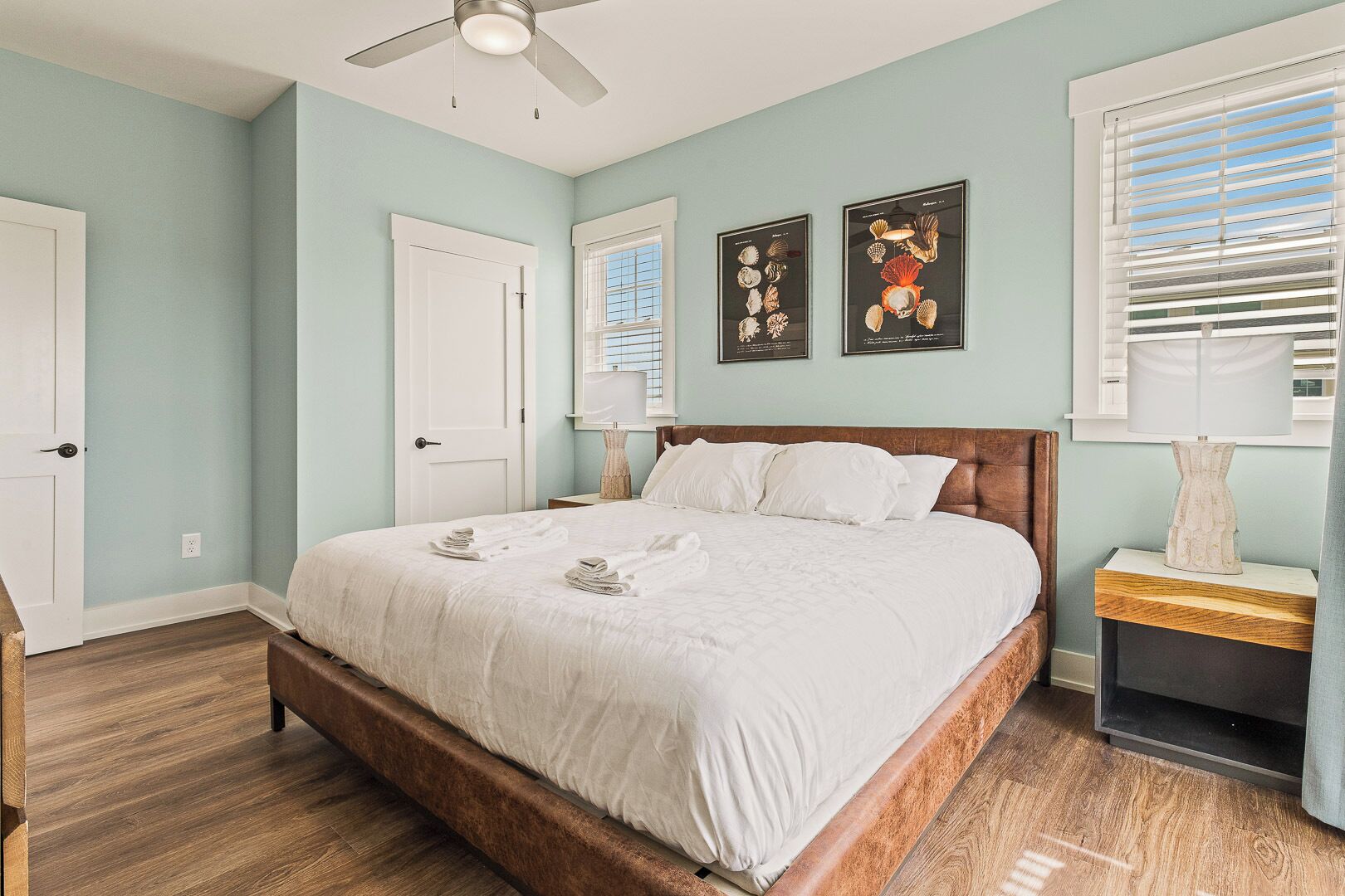 A bedroom with a wooden bed, white bedding, a ceiling fan, a window with blinds, and two framed pictures on the wall.