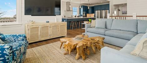 A spacious living room with a large sofa, a coffee table, and a TV. There is a kitchen area with a bar and stools.
