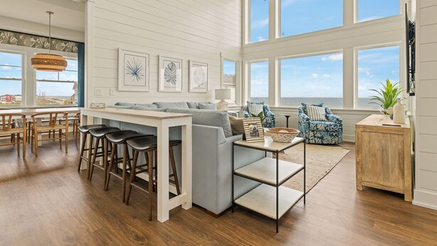 A spacious living room with a large window overlooking the ocean, a dining area with a wooden table and bar stools, and a cozy seating area with patterned chairs.
