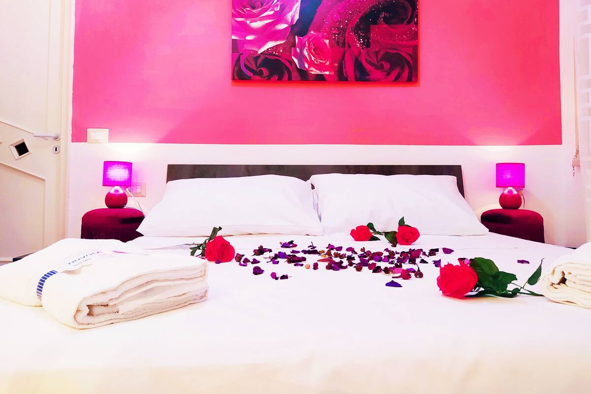 A hotel room with a pink wall, a bed with white linens, two bedside lamps, and a painting above the bed.