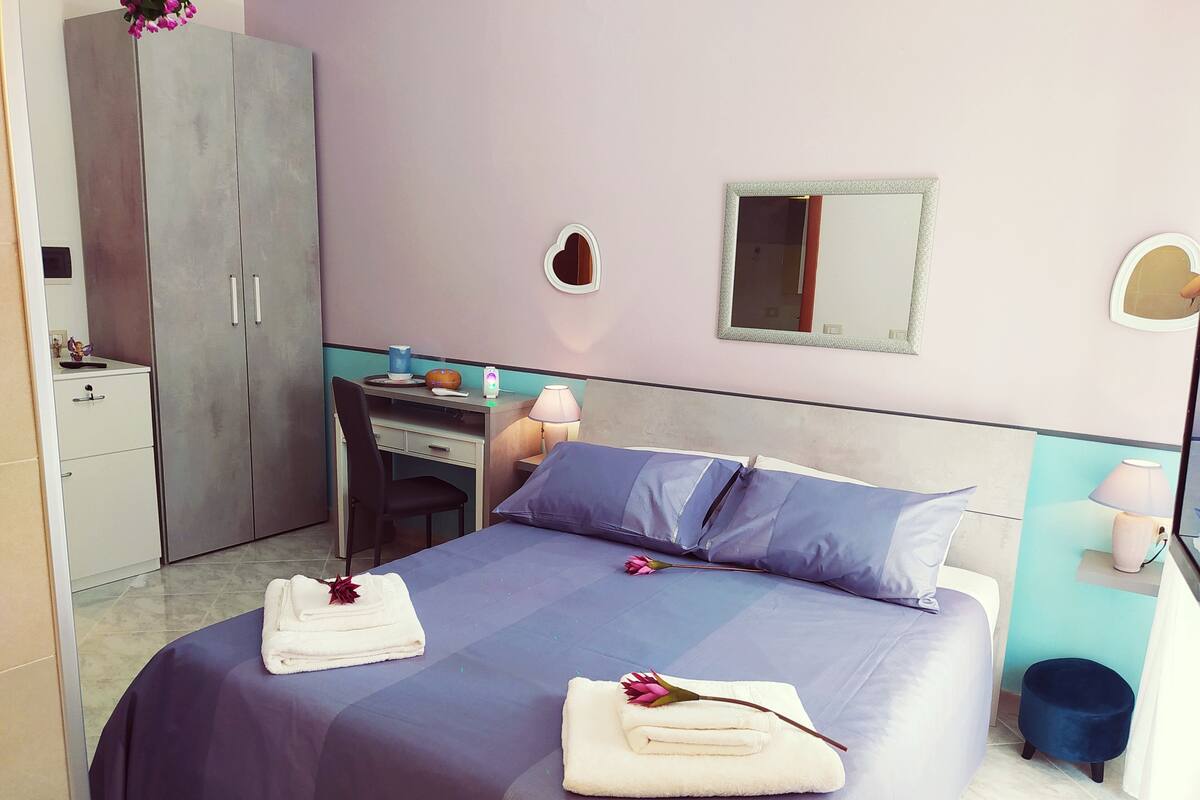 A hotel room with a bed, a desk, a chair, a wardrobe, and a small stool.