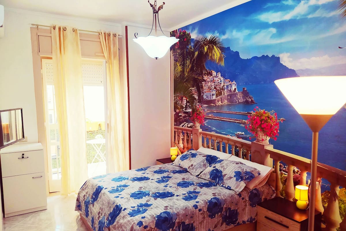 A bedroom with a bed, a nightstand, a lamp, a dresser, and a view of a coastal town through a large window.