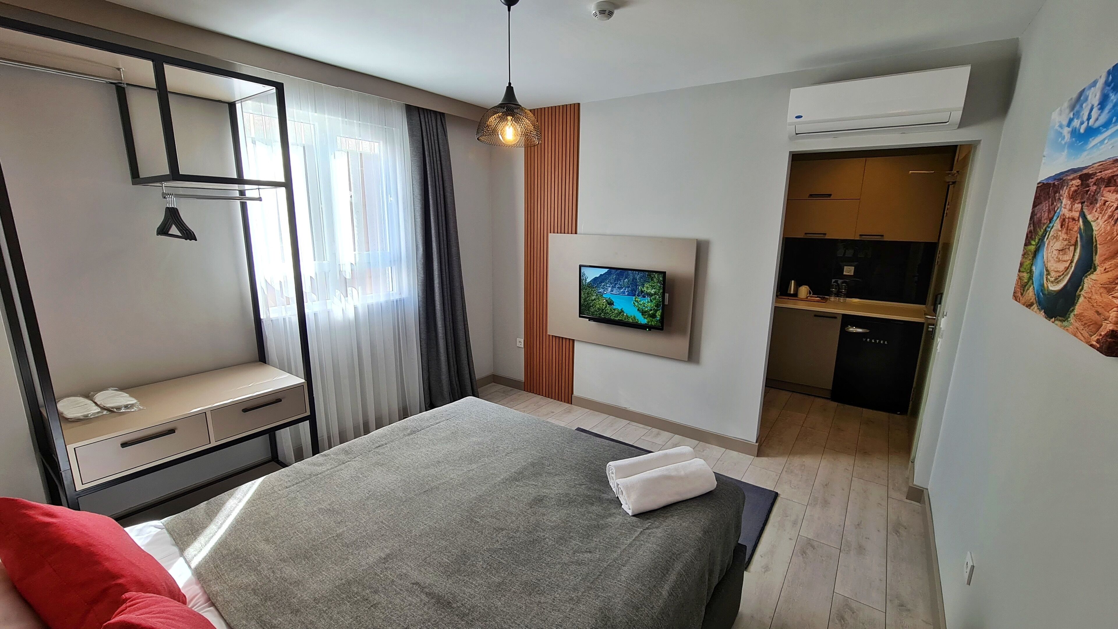 A modern hotel room with a large bed, a TV mounted on the wall, a kitchenette, and a bathroom visible through an open door.