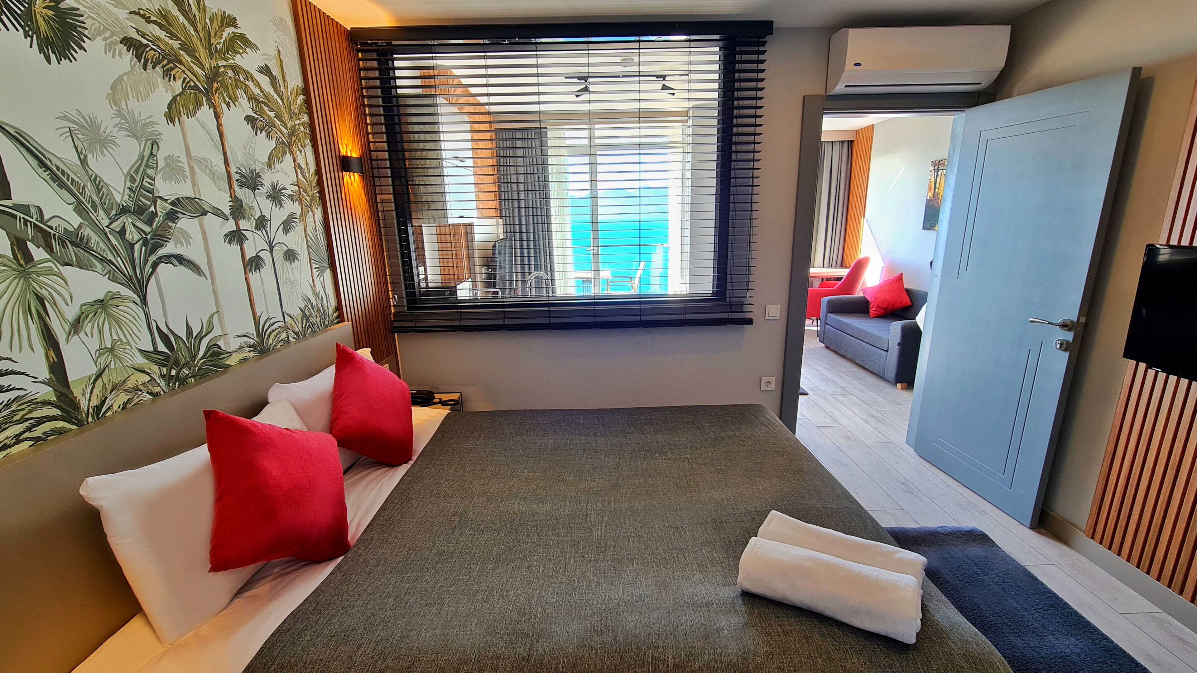 A hotel room with a bed, a window with blinds, a wall with a tropical mural, and a door.