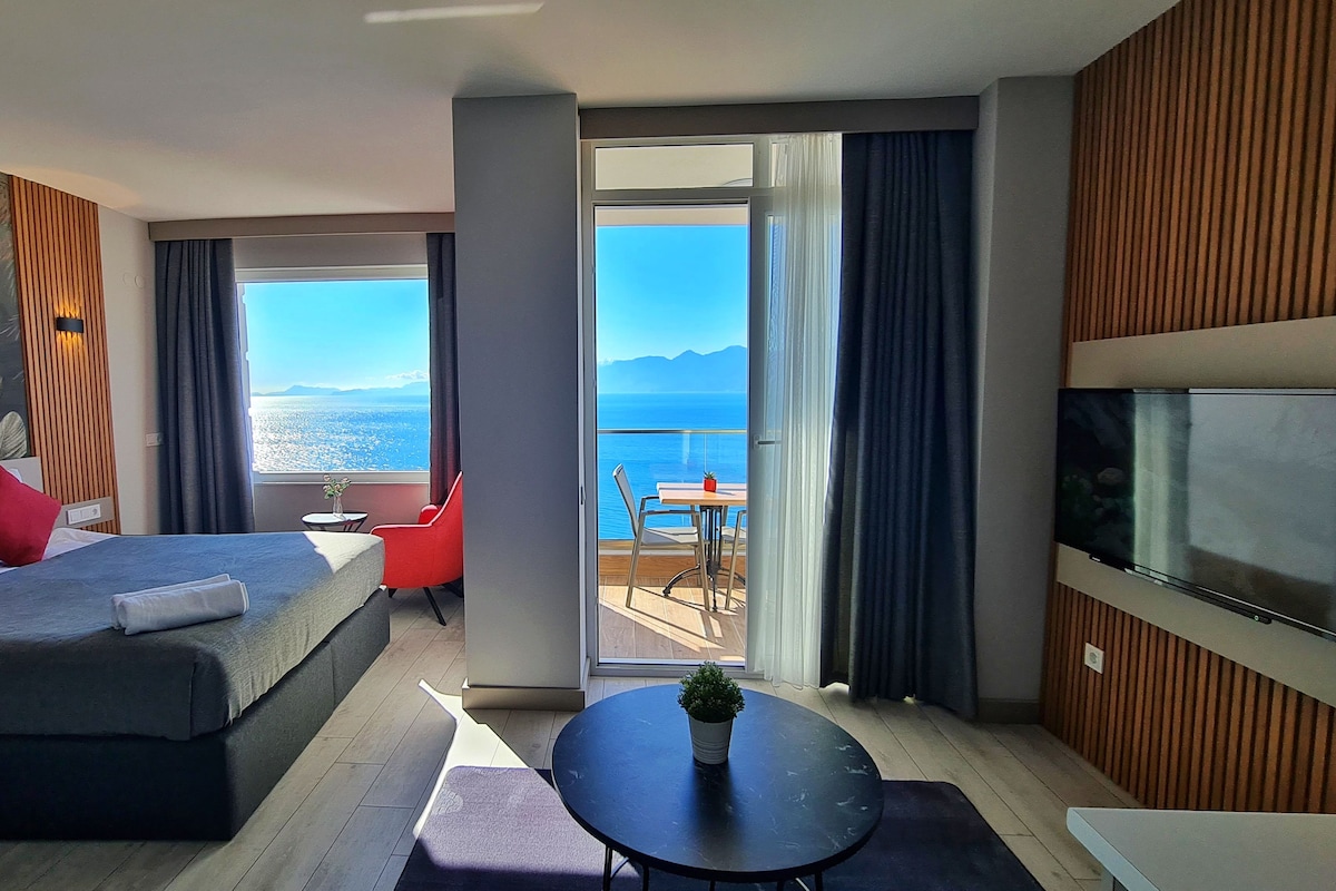 A modern hotel room with a large bed, a TV, a balcony with a view of the sea, and a small table with a plant.