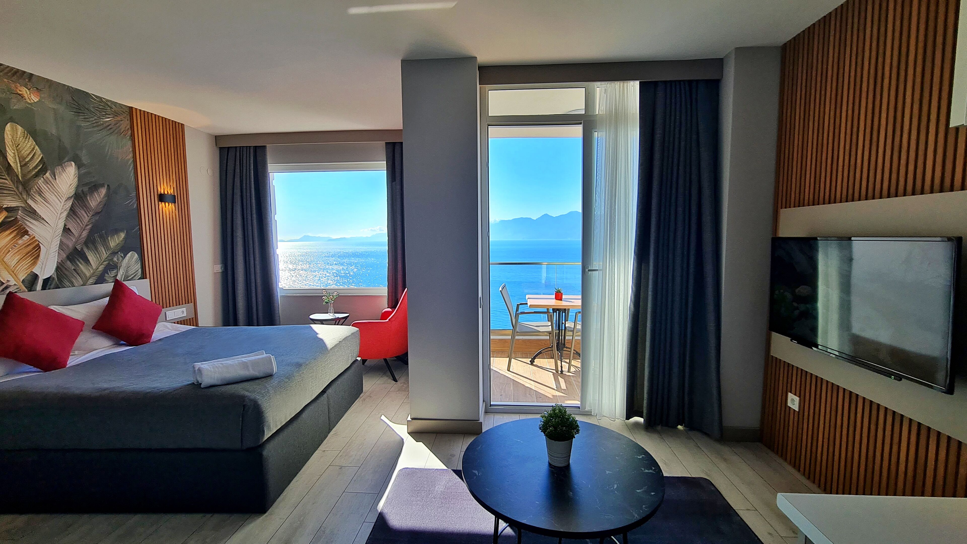 Una habitación de hotel moderna con una cama grande, un sofá, un televisor, un balcón con vista al mar y una mesita con una planta.