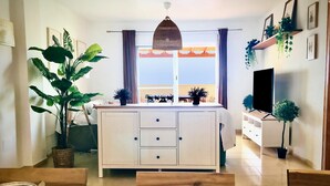 A modern living room with a large wooden dining table, a white sofa, a TV on a stand, and a large window with curtains. - Spectacular apartment with sea views. WIFI. SmartTV 58” (Los Geraneos)