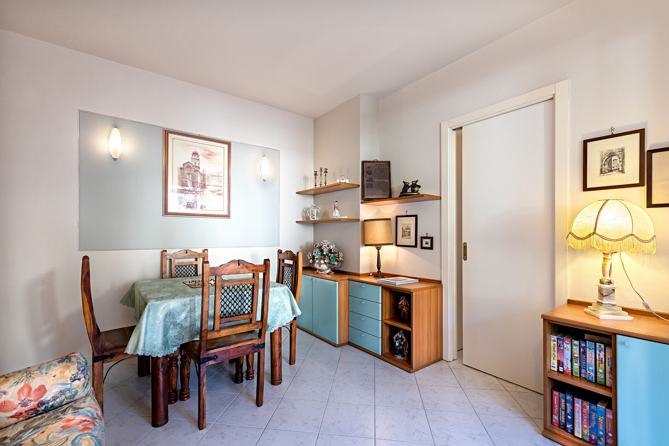 A dining area with a table set for two, wooden chairs, a cabinet with drawers, and a bookshelf with books.