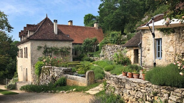 Une maison en pierre avec un toit recouvert de tuiles rouges, entourée d’une végétation luxuriante et de plantes en pot.
