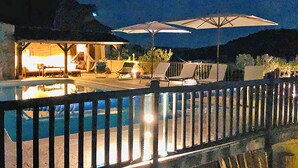 Une vue nocturne d’une piscine, avec des chaises longues et des parasols, une terrasse en bois et un bâtiment illuminé.