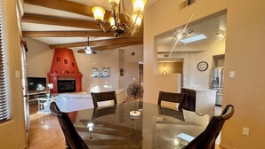 A dining area with a glass table and wooden chairs, a fireplace, and a kitchen in the background. - Sage Swan, 30+ Day Rentals, Near I-40 (Albuquerque)