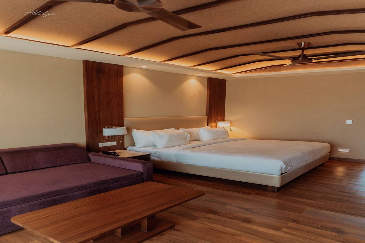 A modern hotel room with a wooden ceiling, a bed with white linens, a purple sofa, and a wooden bench.