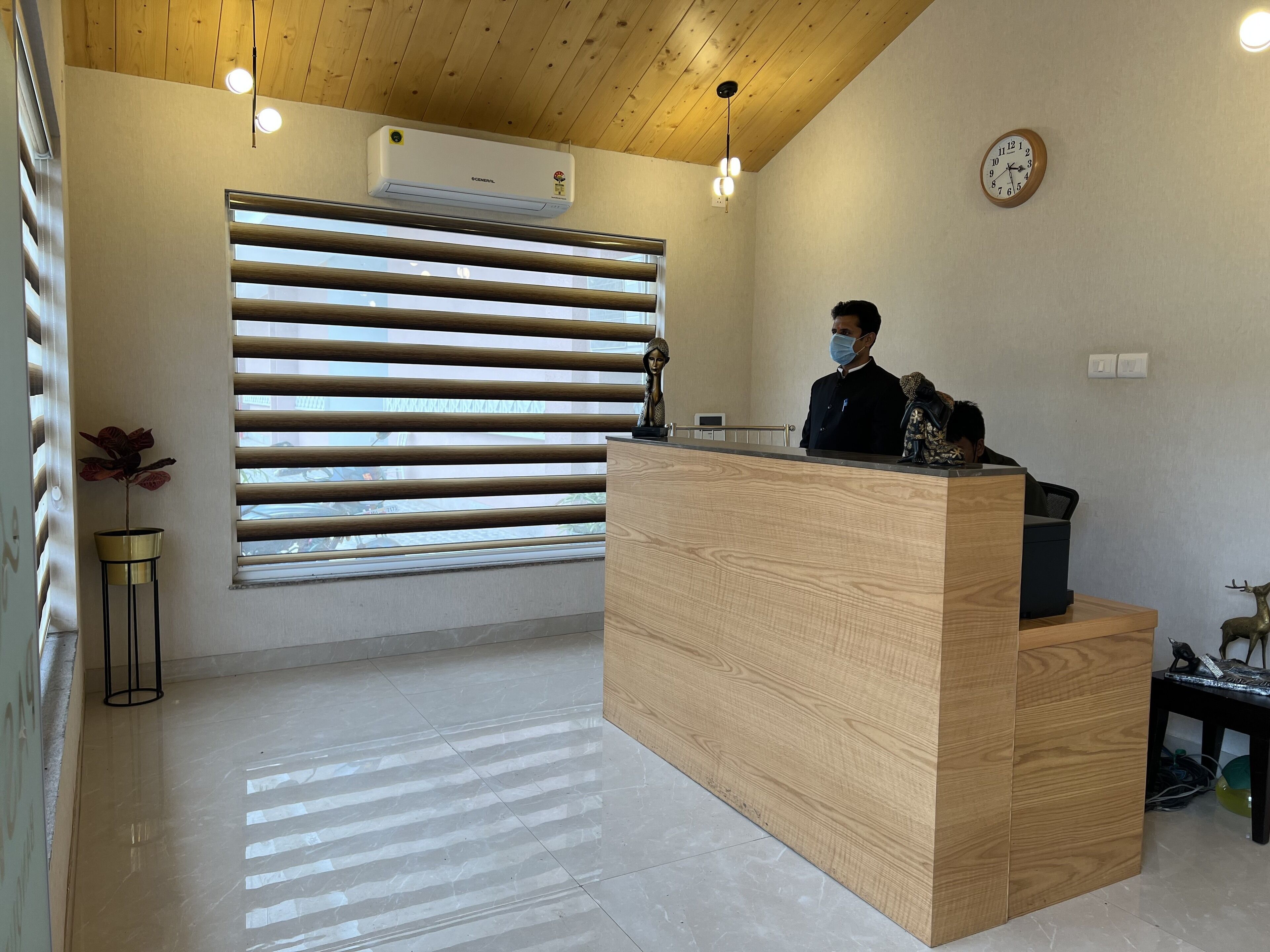 Un mostrador de recepción con detalles de madera, un reloj en la pared y una ventana con persianas horizontales.