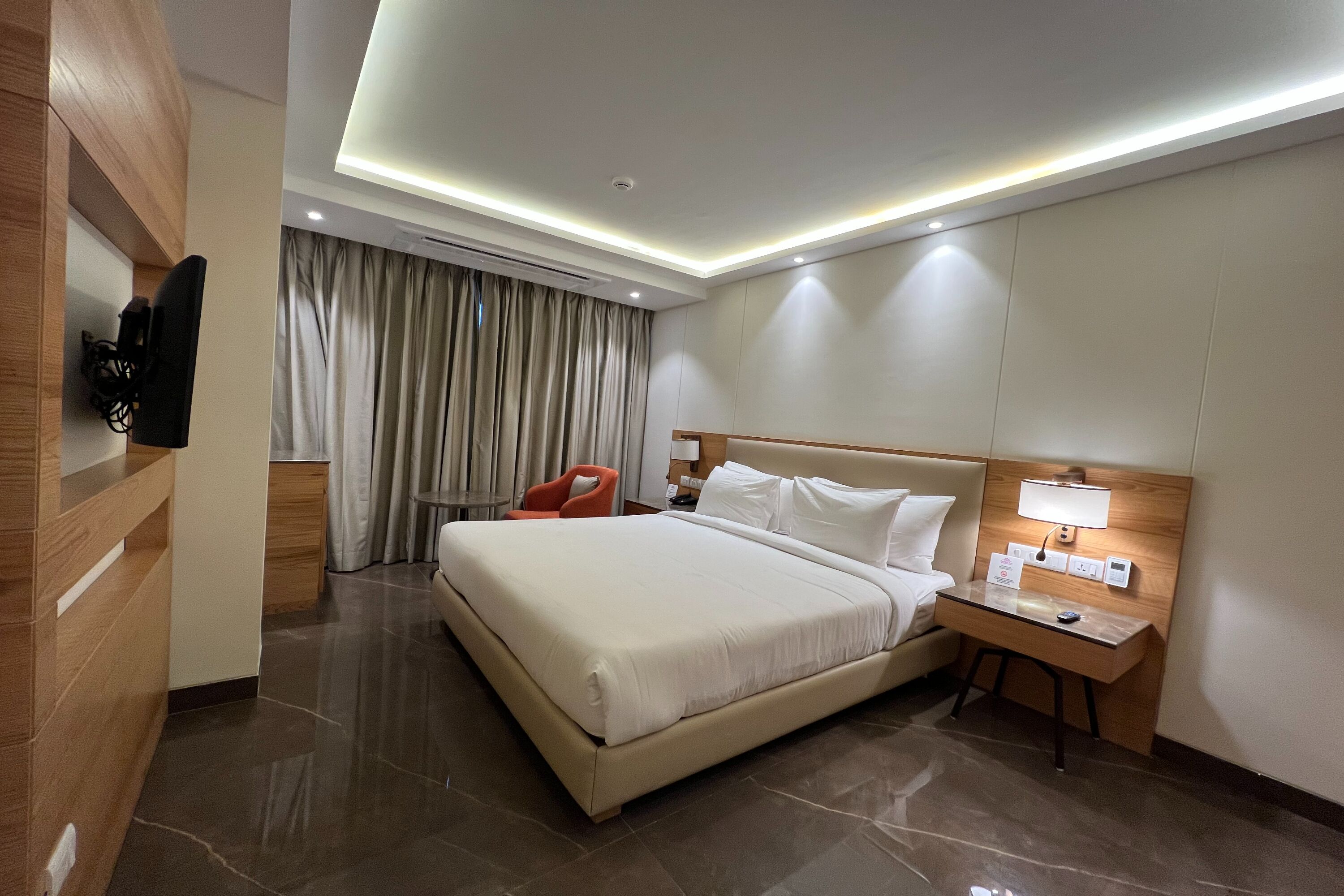 Habitación de hotel con una cama grande, un escritorio con lámpara, un televisor montado en la pared y una ventana con cortinas.