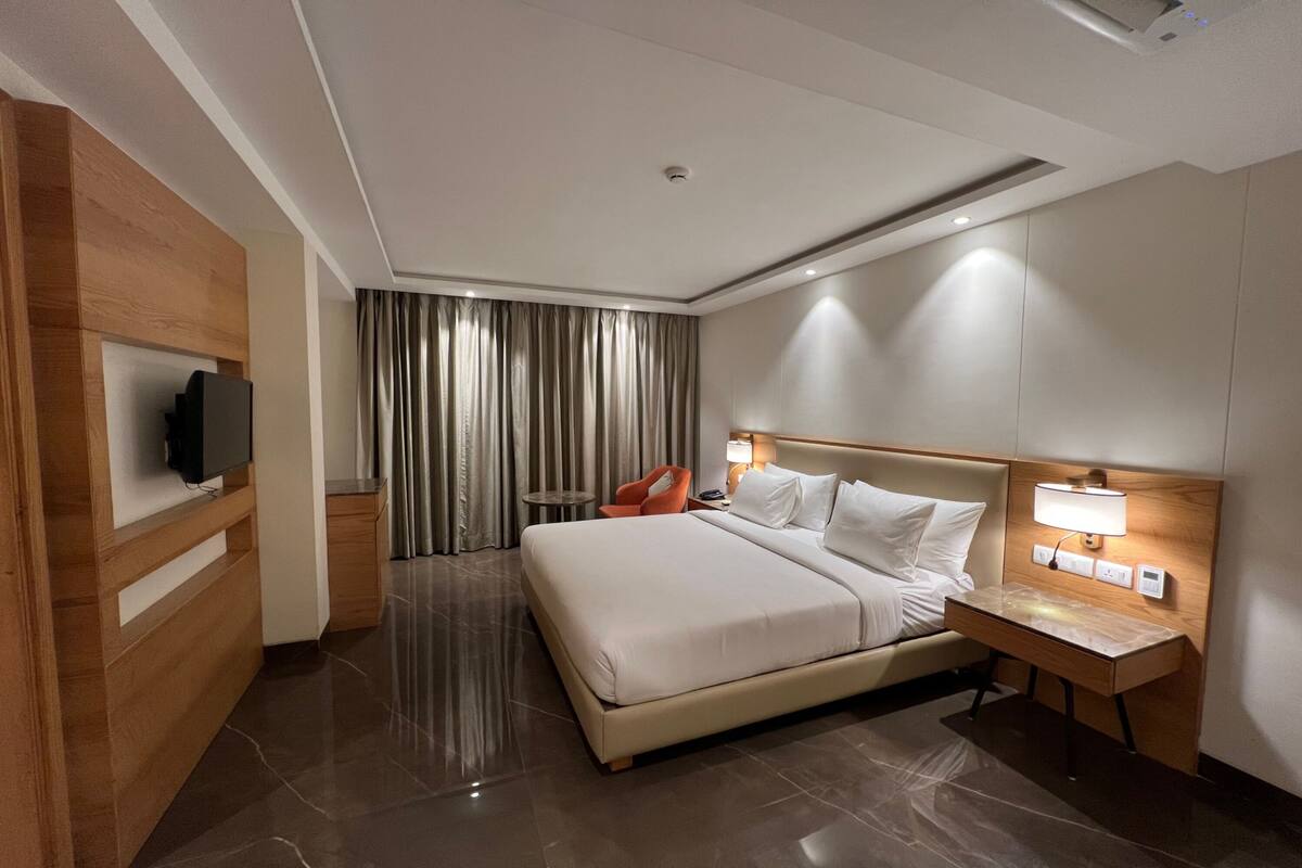 A modern hotel room with a large bed, a bedside table with a lamp, a TV mounted on the wall, and a small table with a chair.