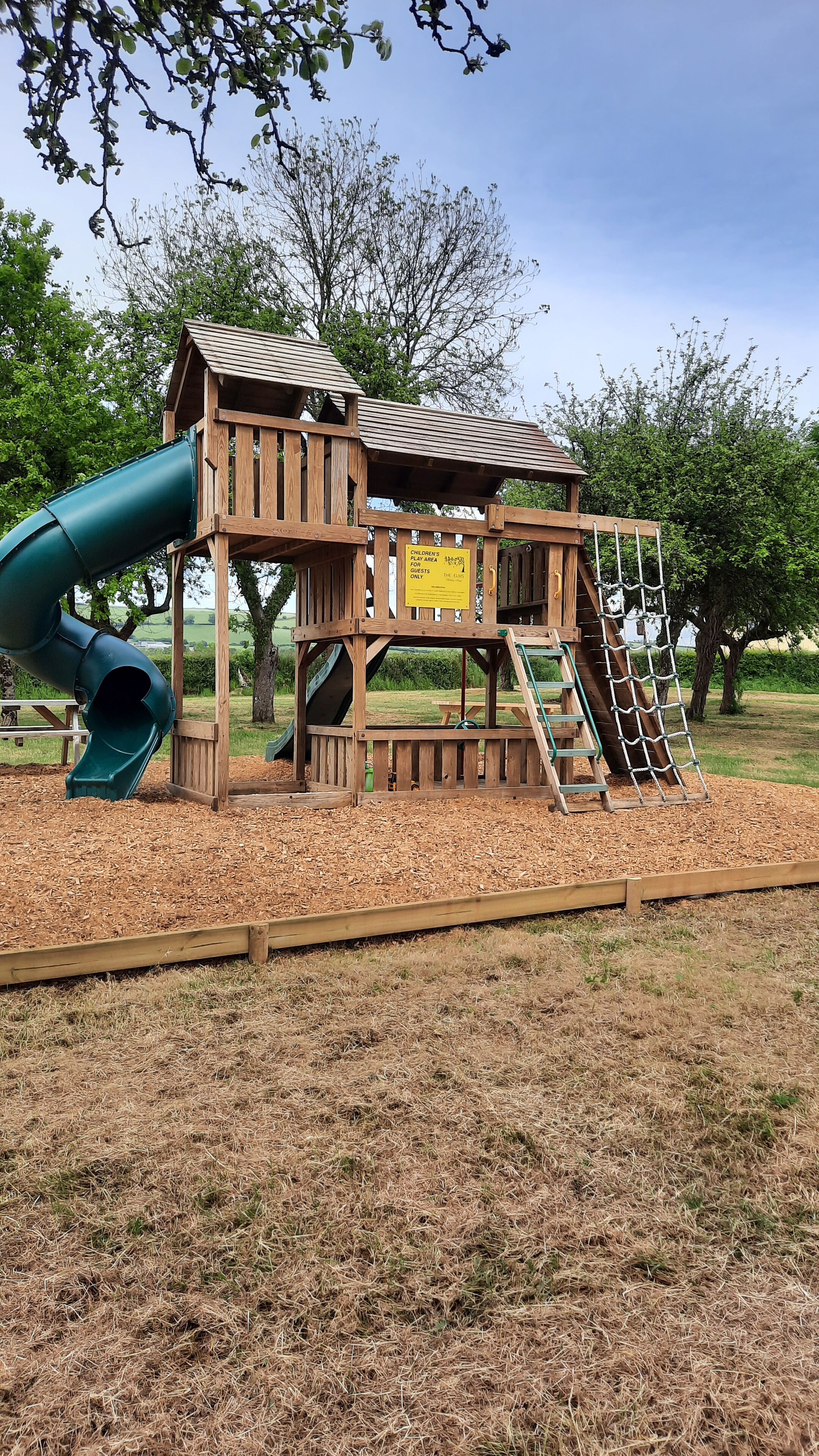 A wooden play structure with a slide and climbing net, surrounded by mulch and trees.
