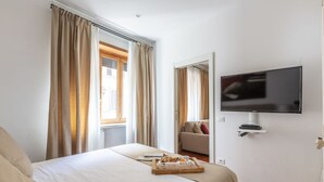 A hotel room with a bed, a flat-screen TV mounted on the wall, a window with curtains, and a wooden side table with a tray of breakfast items. - 2 Apartments same building Piazza del Popolo 1+3 (Roma)