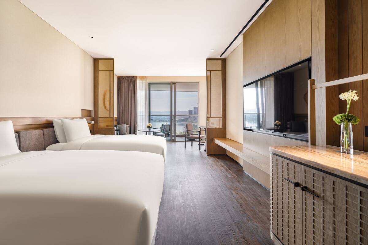 A hotel room with two beds, a TV, a desk, and a view of the city.