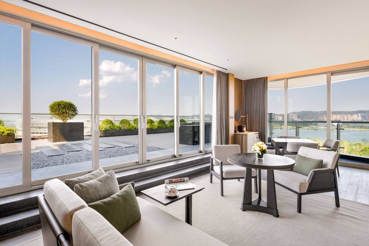 A modern hotel room with large windows, a sofa, a table, and a chair.