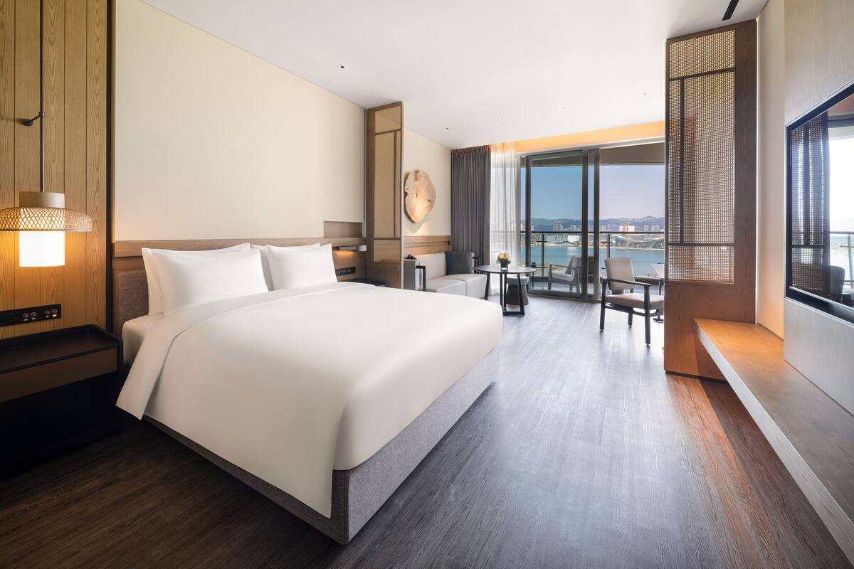 A modern hotel room with a large bed, a desk, and a balcony with a view.
