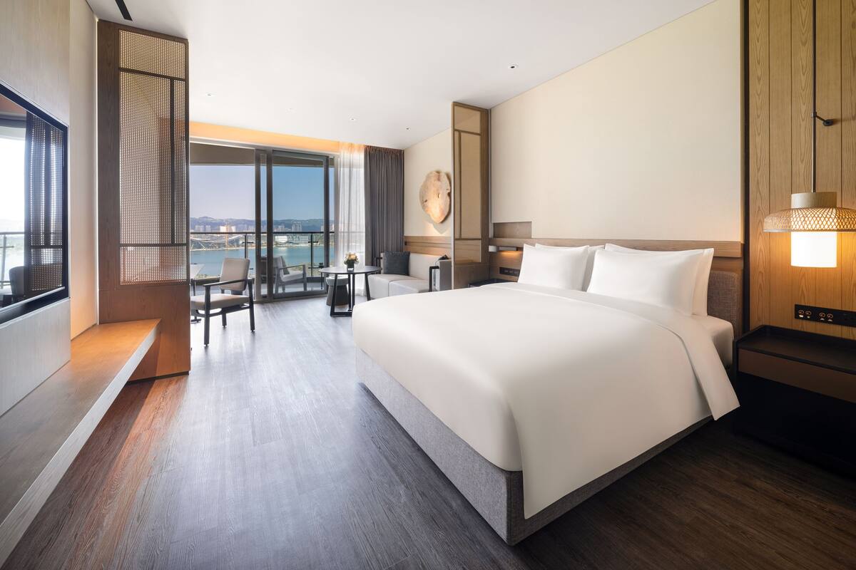 A modern hotel room with a large bed, a balcony with a view, a desk, and a seating area.