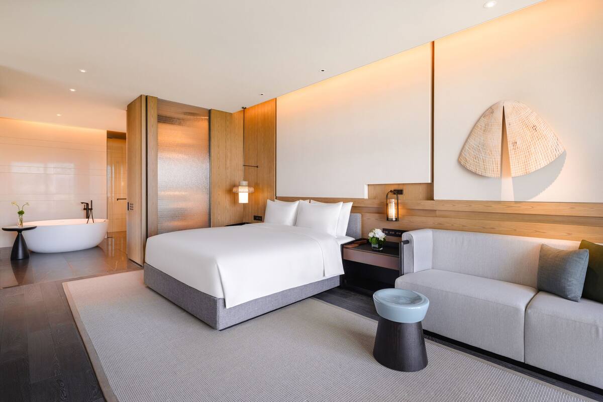 A modern hotel room with a large bed, a sofa, a small table, and a bathtub.
