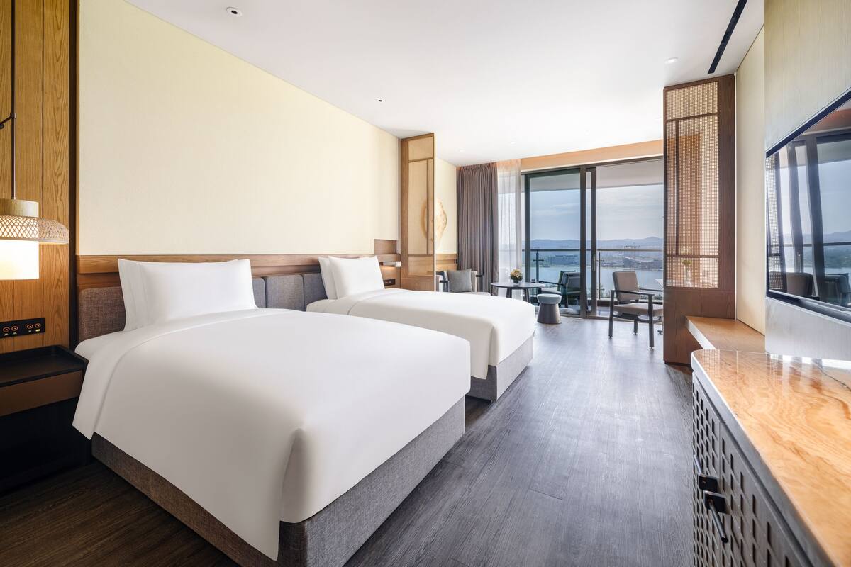 A hotel room with two beds, a desk, a chair, and a large window offering a view of the city.