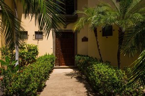 A pathway leading to a building with a wooden door, surrounded by lush greenery and palm trees.