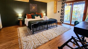 A bedroom with a wooden ceiling, a bed with multiple pillows, a bedside table with a lamp, and a view of the outdoors through a window.