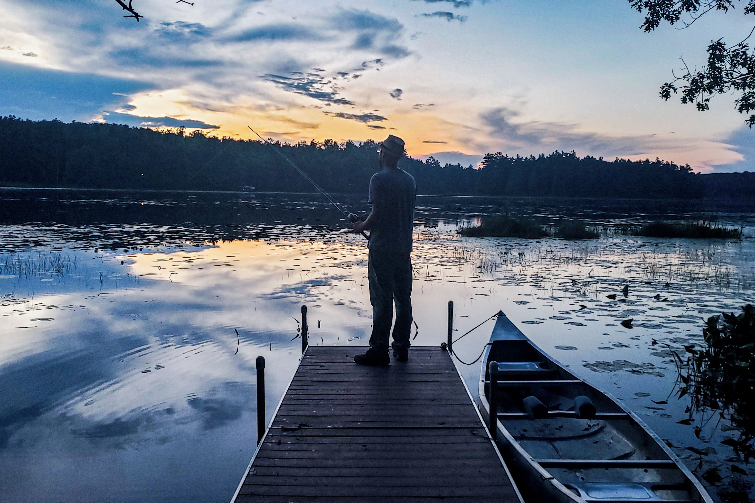 A person standing on a wooden dock extending into a calm body of water, with a boat moored at the dock's end.