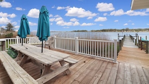 A wooden deck with picnic tables and umbrellas, overlooking a calm body of water and a distant shoreline. - An Unfettered View | Wheelchair Accessible Oyster Bay Waterfront Ranch w/ Long Pier for Crabbing (Chincoteague Island)