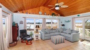A spacious living room with a wooden ceiling, a grey sectional sofa, a black leather chair, a patterned ottoman, and a view of the outdoors through large windows. - An Unfettered View | Wheelchair Accessible Oyster Bay Waterfront Ranch w/ Long Pier for Crabbing (Chincoteague Island)