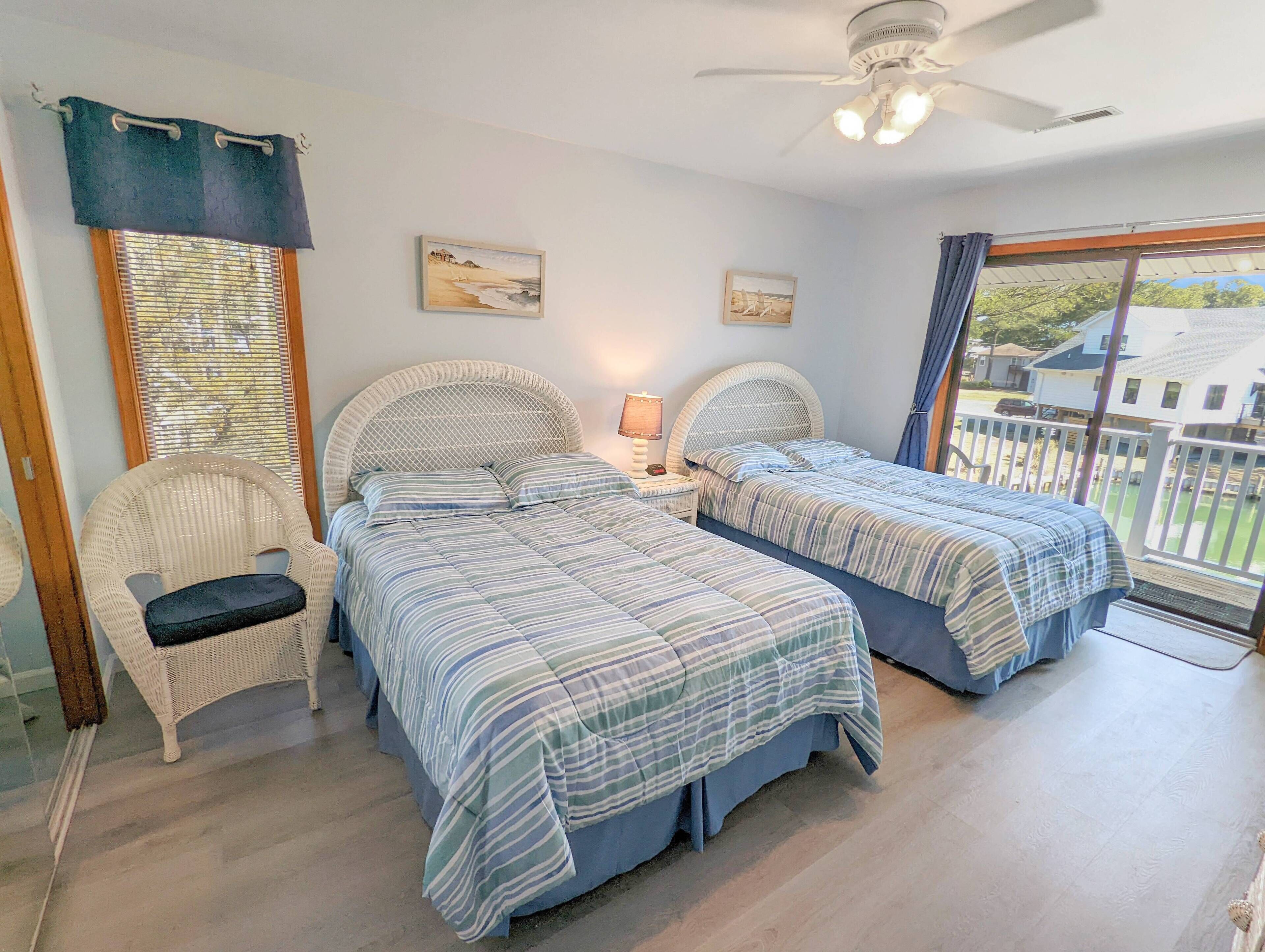 A bedroom with two beds, a chair, a ceiling fan, and a window with a view of a house.