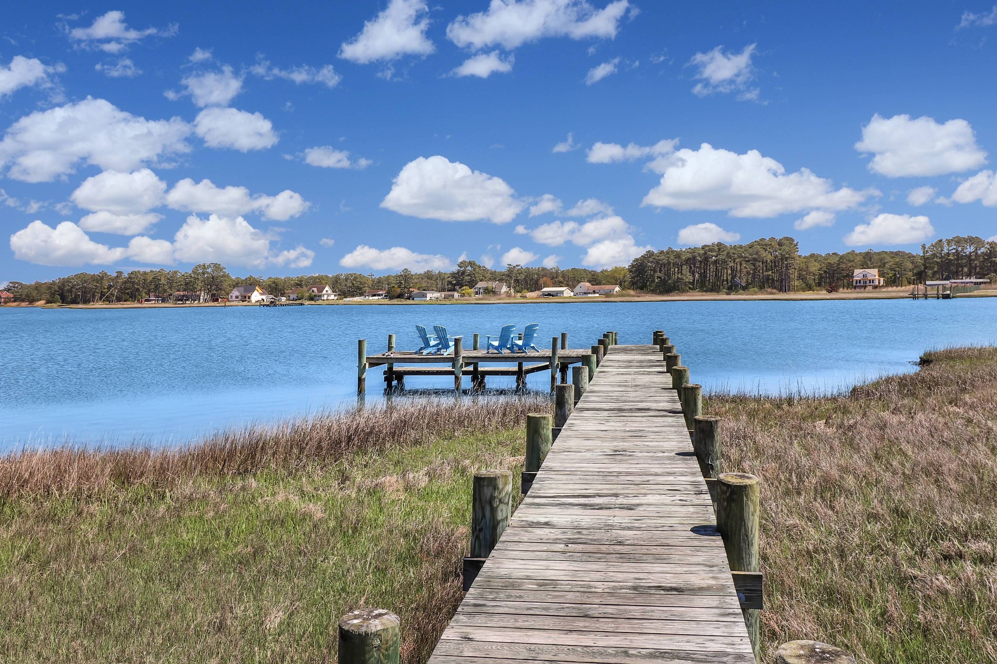 Eine hölzerne Mole, die sich in ruhiges Wasser erstreckt, mit blauem Himmel und Häusern in der Ferne.