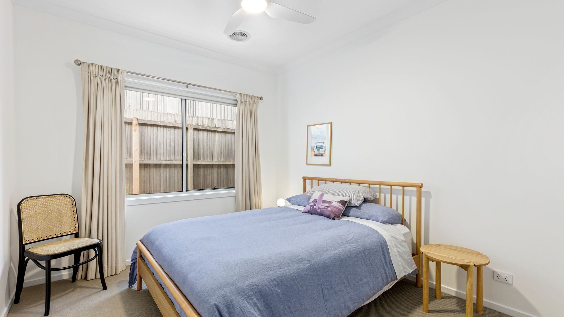 A bedroom with a bed, a chair, a small table, a window with curtains, and a ceiling fan.
