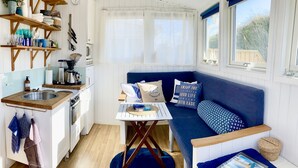 A compact kitchen with a sink, stove, and wooden shelves. A blue sofa with patterned cushions is against the wall, and a small table with a basket and a tablet is in the corner. - Beach cottage No. 7 SKREA STRAND (Hallands län)