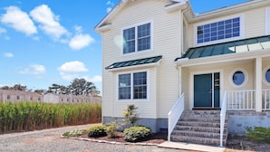 A two-story house with a front porch, a green door, and a small front yard with plants. - Seas the Day | Bright Duplex + Screened Porch + Community Pool + Kid Bunk Room (Chincoteague Island)