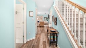 A hallway with a wooden floor, a staircase with white balusters, and a wall-mounted shelf with a bowl and cans. - Seas the Day | Bright Duplex + Screened Porch + Community Pool + Kid Bunk Room (Chincoteague Island)