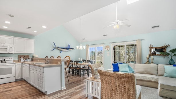 A modern kitchen with white cabinets, a dining area with wooden chairs, a wicker sofa, and a ceiling fan. - Seas the Day | Bright Duplex + Screened Porch + Community Pool + Kid Bunk Room (Chincoteague Island)