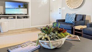 A living room with a television displaying a beach scene, a sofa, a coffee table with a succulent plant, and a book titled "Chicacara Island". - Serenity at Oyster Bay | Screened Porch, Preserve Views + Near Water Access, Firepit (Chincoteague Island)