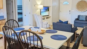 A modern living room with a dining area, featuring a large table, chairs, a ceiling fan, and a TV displaying a beach scene. - Serenity at Oyster Bay | Screened Porch, Preserve Views + Near Water Access, Firepit (Chincoteague Island)