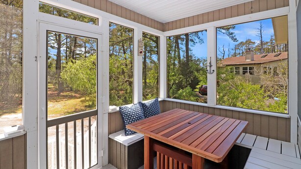 A covered patio with a wooden table and bench, large windows offering a view of trees and a house. - Serenity at Oyster Bay | Screened Porch, Preserve Views + Near Water Access, Firepit (Chincoteague Island)