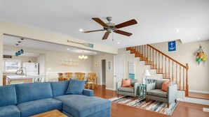 A spacious living room with a blue sectional sofa, a wooden coffee table, and a staircase with a wooden railing. - Shell Cottage | East Side Assateague Channel Views w/ Decks, Outdoor Shower, Boat Parking & Pets (Chincoteague Island)