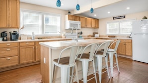 A modern kitchen with wooden cabinets, a white island with bar stools, and pendant lights. - Shell Cottage | East Side Assateague Channel Views w/ Decks, Outdoor Shower, Boat Parking & Pets (Chincoteague Island)
